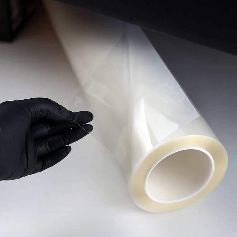 Gloved hand peeling Feynlab PPF Flex clear film from the roll showing 7.5 mil thickness and optical clarity
